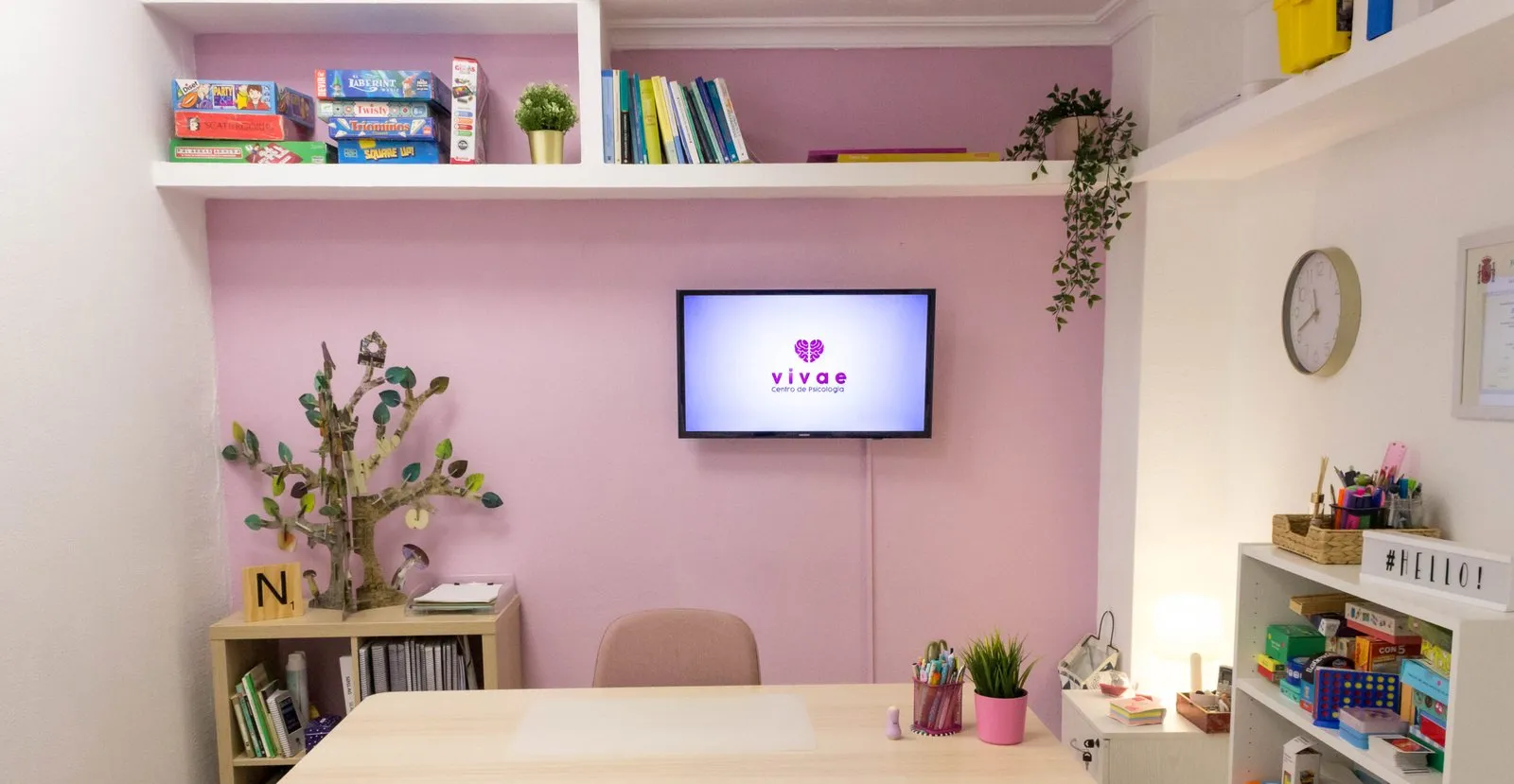 Despacho de pared rosa con mesa de madera, estantería con juegos y libros y pantalla con el logo de Vivae